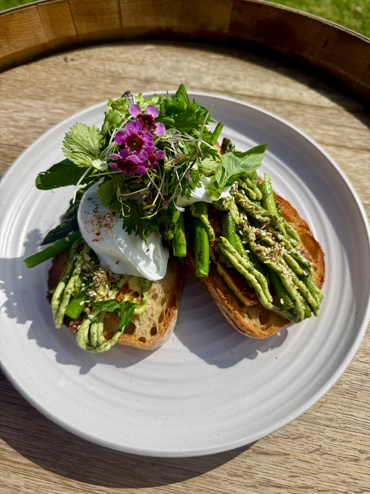 Two slices of avocado toast on a white plate, topped with poached egg, microgreens, and edible flowers, set on a wooden table outdoors.