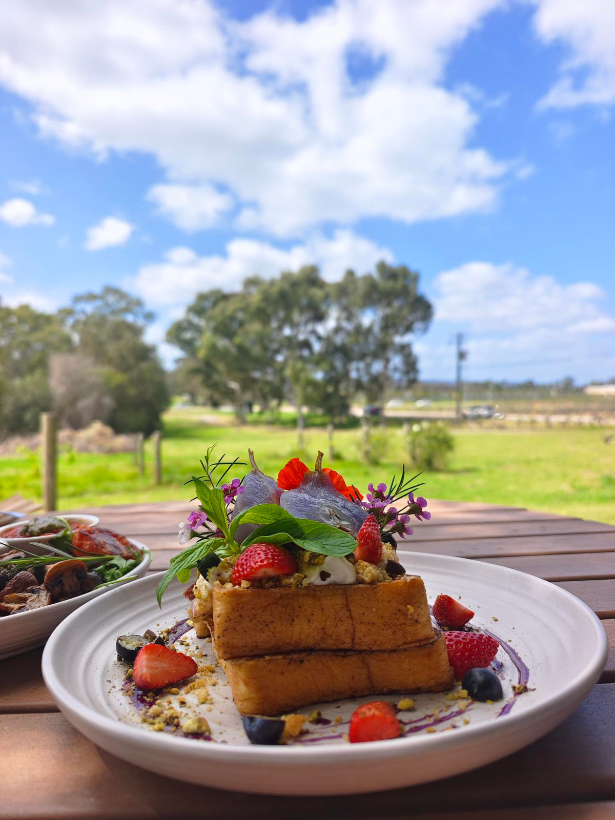 A plated stack of golden-brown French toast or pancakes topped with fresh strawberries, blueberries, purple flowers, and green leaves, set on a wooden table outdoors with a blurred field and blue sky in the background.