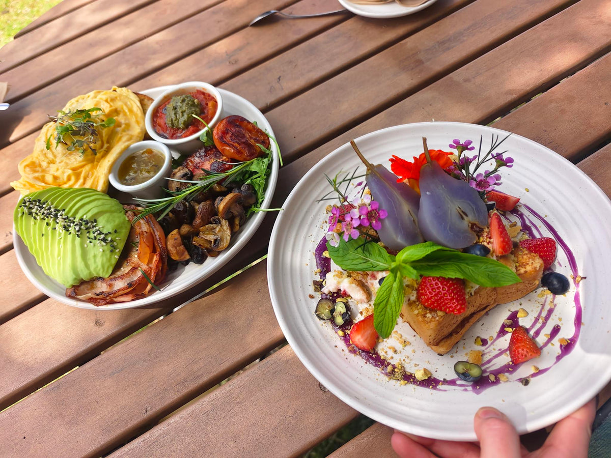 Two plated breakfast dishes on a wooden outdoor table. One plate contains an omelette, avocado, bacon, mushrooms, and small bowls of sauce.
