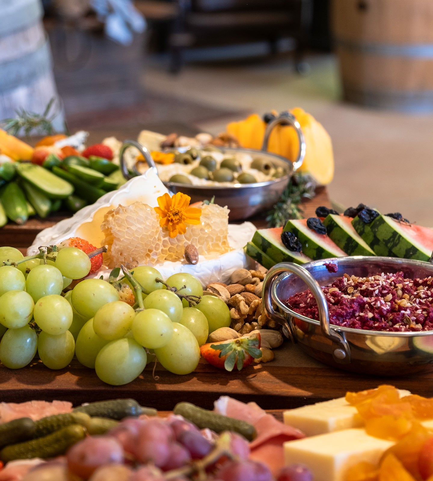 A rustic charcuterie or cheese board arranged on a wooden table, featuring green grapes, sliced watermelon, cucumber, nuts, honeycomb, crackers, and bowls of appetizers in a warm indoor setting.