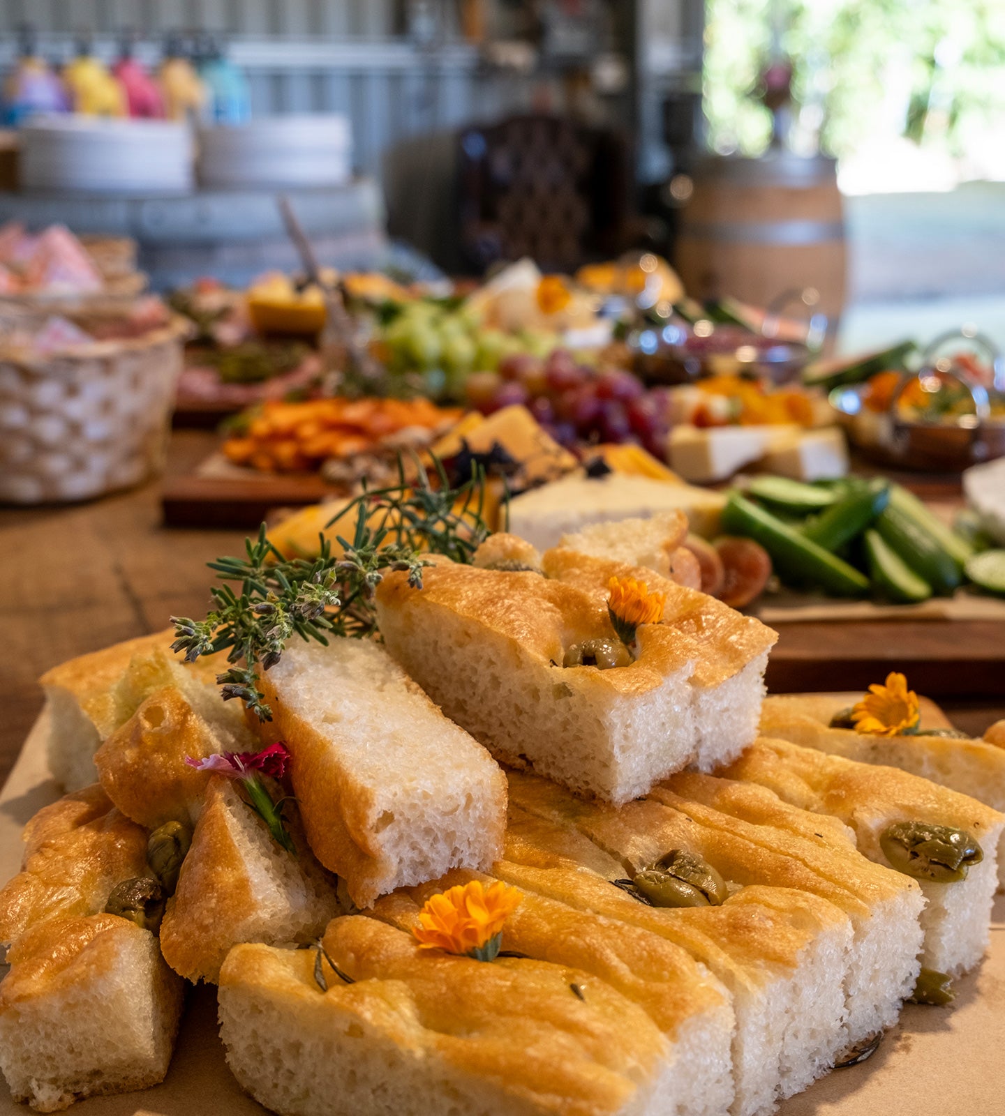 Close-up of freshly baked focaccia bread sticks topped with herbs and edible flowers, arranged on a wooden board in a rustic restaurant setting with charcuterie boards blurred in the background.