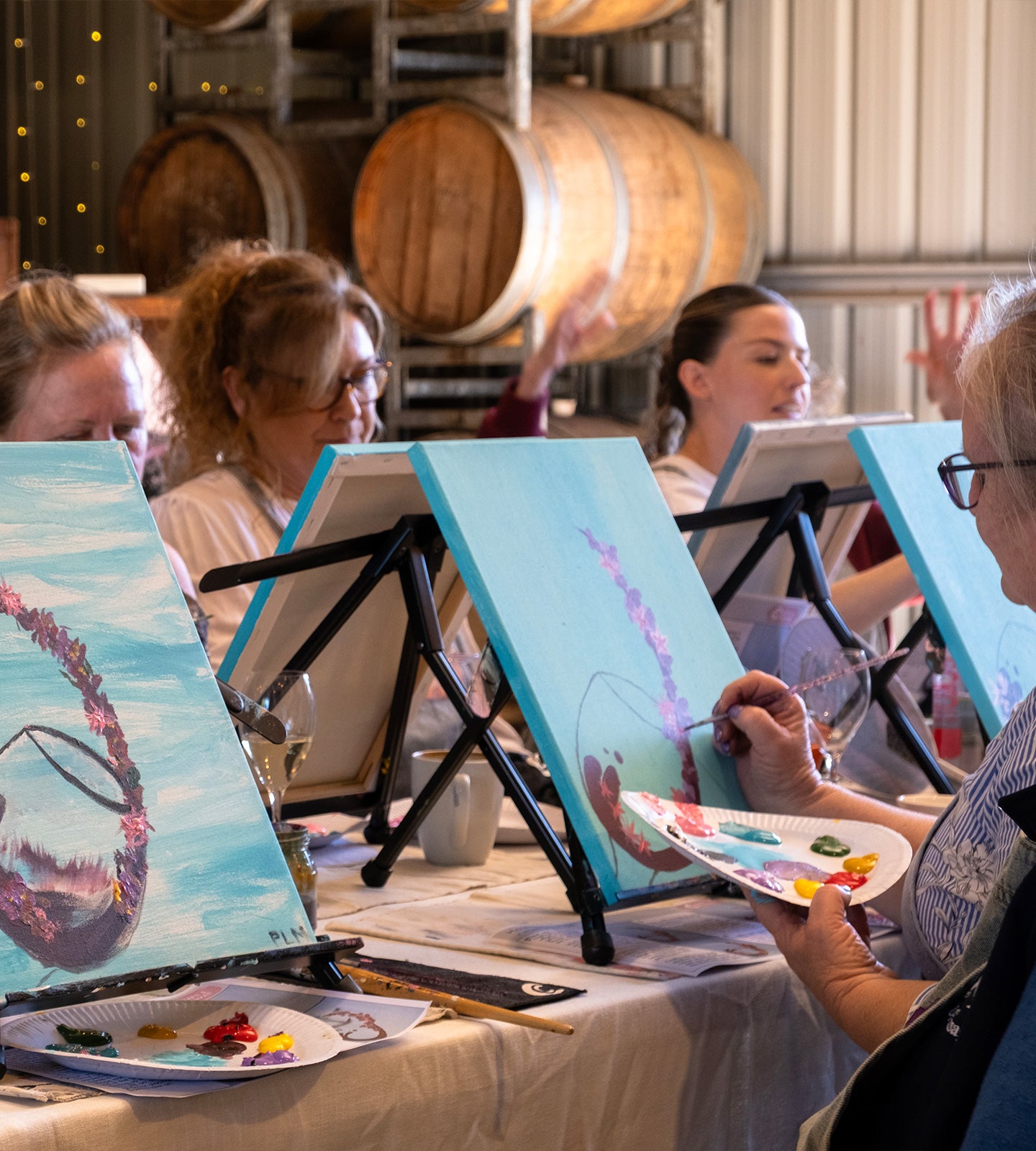 Several people painting at easels in a workshop or studio, with wooden barrels visible in the background. One person in the foreground holds a paint palette.
