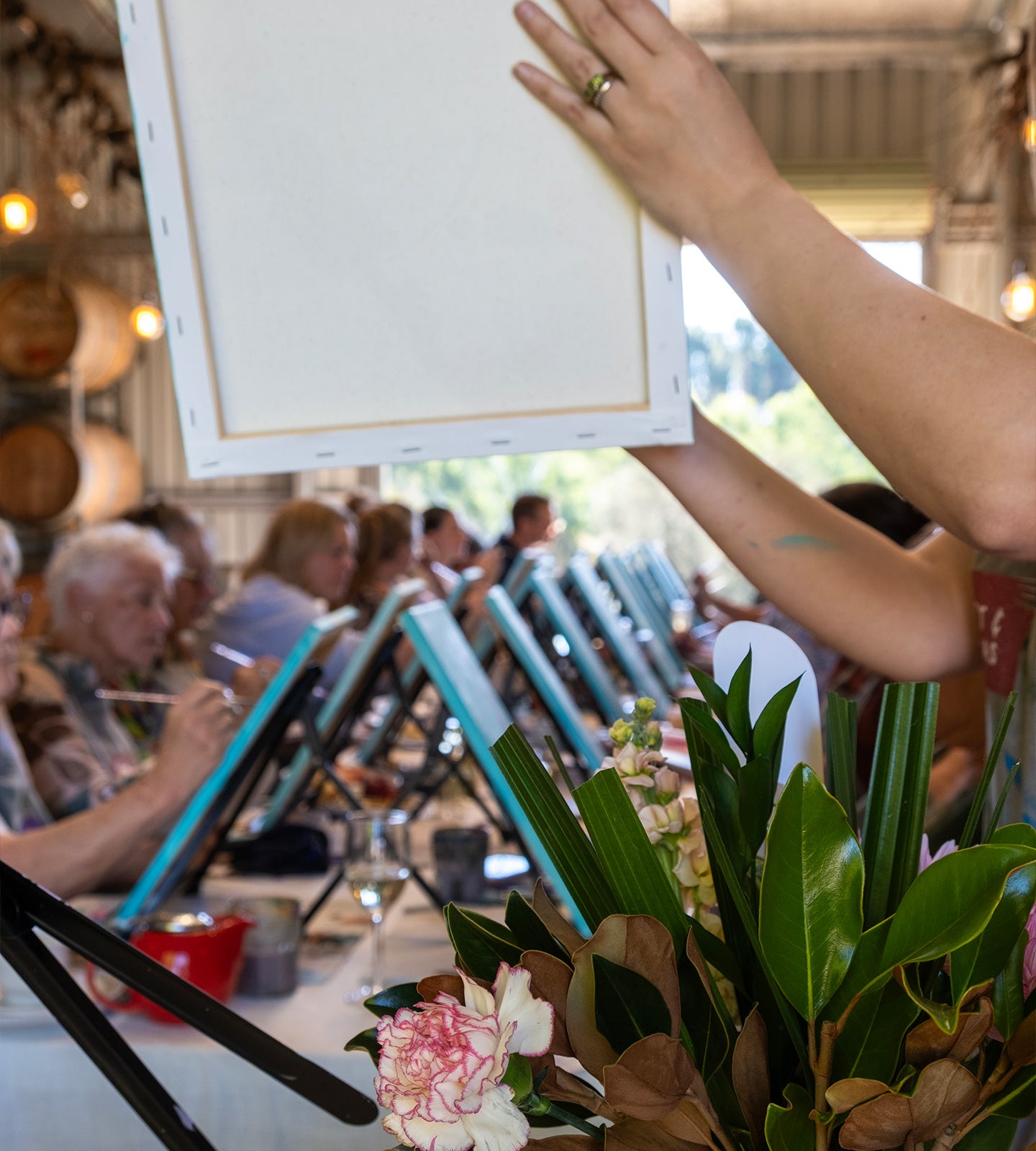 A person at a table holding a blank canvas in a painting class, with other participants and floral arrangements blurred in the background.