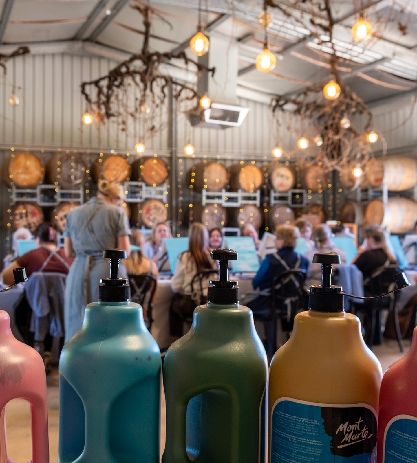 A row of colorful pump bottles in the foreground, with a blurred indoor event or workshop setting in the background where people are seated and standing.