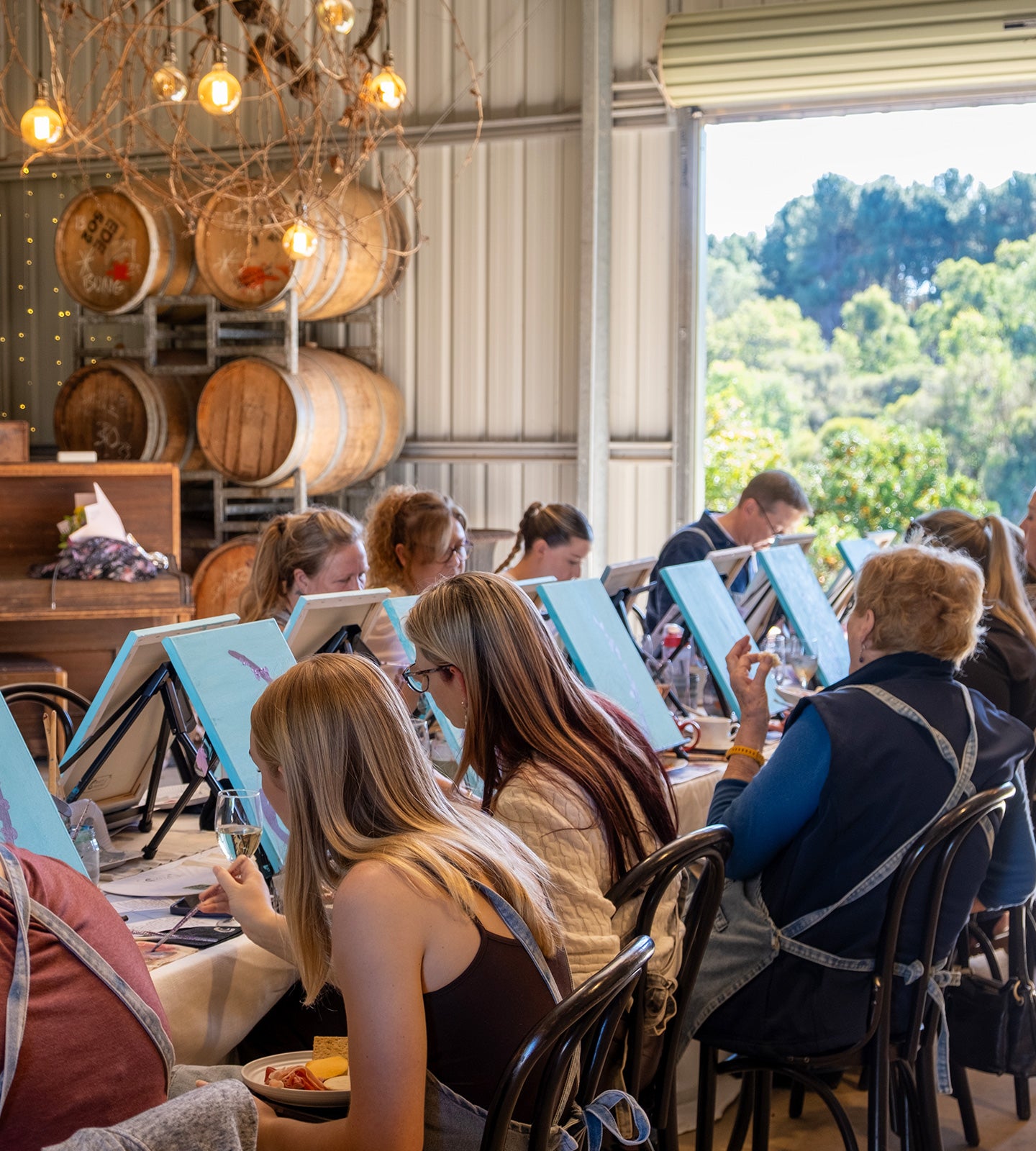A group of people seated indoors at tables with blue-framed artboards or sketchbooks, in a rustic room with exposed wood walls, hanging lights, and barrels. A large window shows green trees outside.