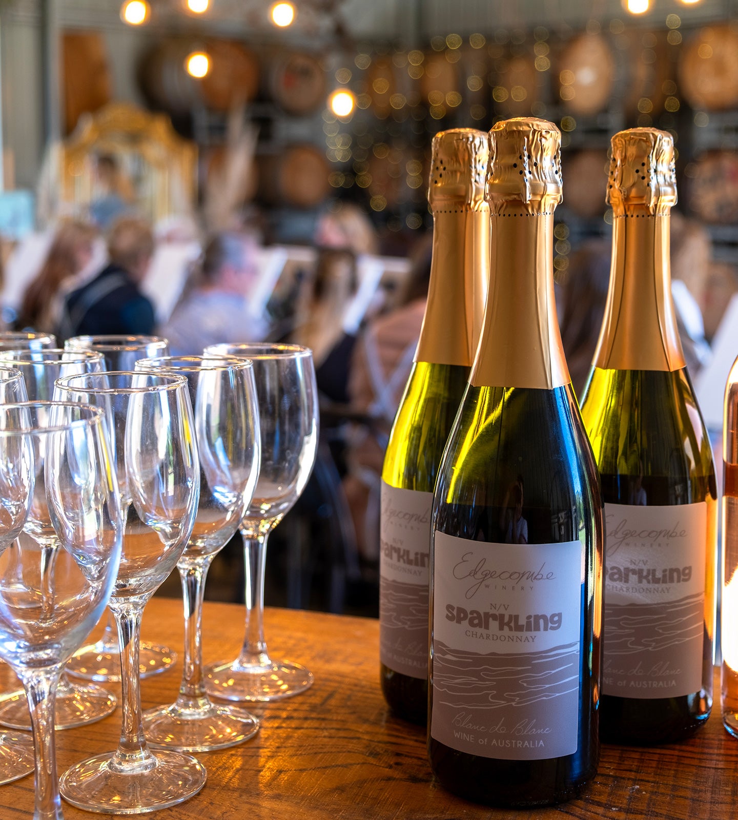 Three sparkling wine bottles with gold foil and white labels on a wooden table, with several clear wine glasses in the foreground and a blurred indoor event background with people and warm lights.